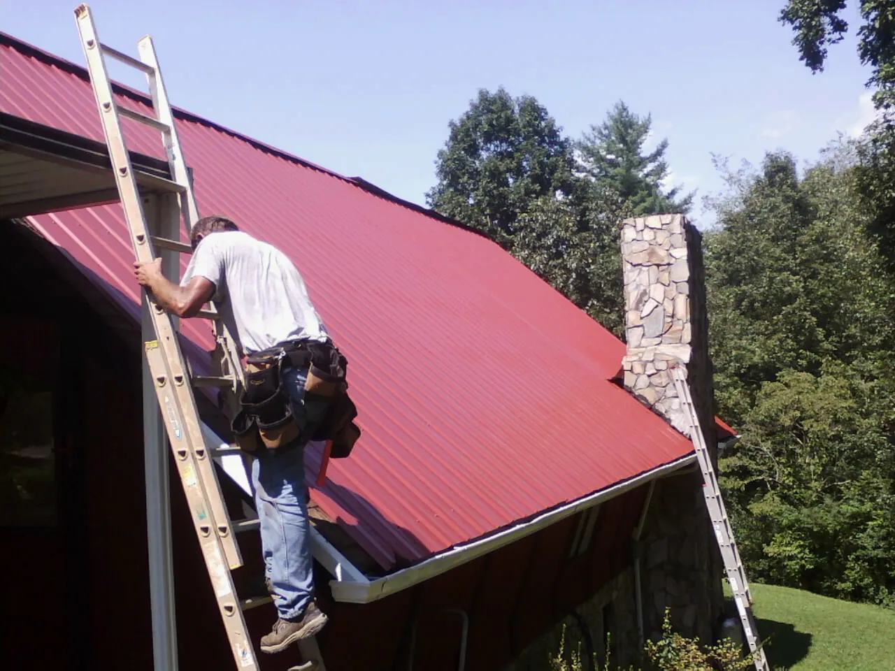 Roofer on a ladder beside a red metal roof and stone chimney — Soffit & Fascia Repair by Roofing Brothers in Mount Hamilton